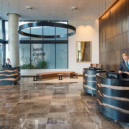 Pullman Brisbane Airport Hotel Exterior photo The image shows a modern hotel lobby. There are two reception desks made of dark-colored materials, and staff members are attending to guests at these desks. The lobby features large windows that allow natural light to fill the space, and there are p