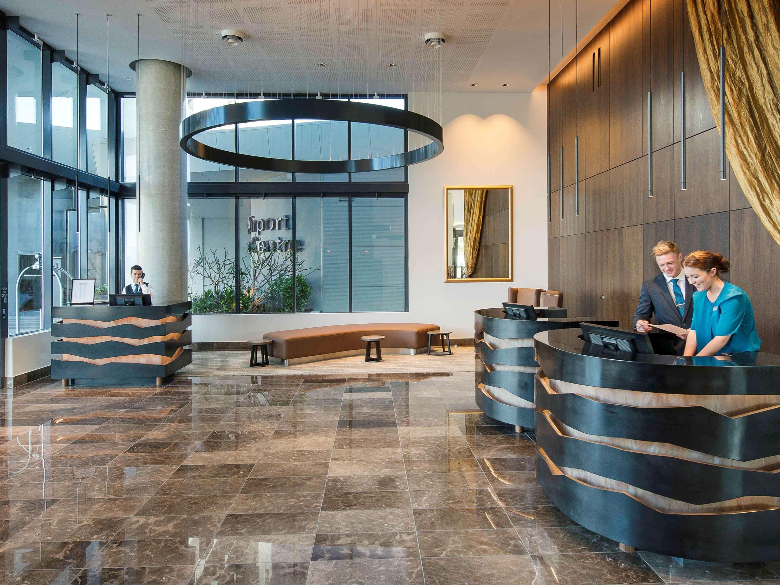 Pullman Brisbane Airport Hotel Exterior photo The image shows a modern hotel lobby. There are two reception desks made of dark-colored materials, and staff members are attending to guests at these desks. The lobby features large windows that allow natural light to fill the space, and there are p